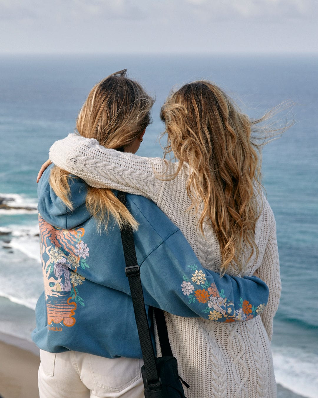 Two women embracing in Saltrock chunky cable knit open front cardigans and looking out at the sea.