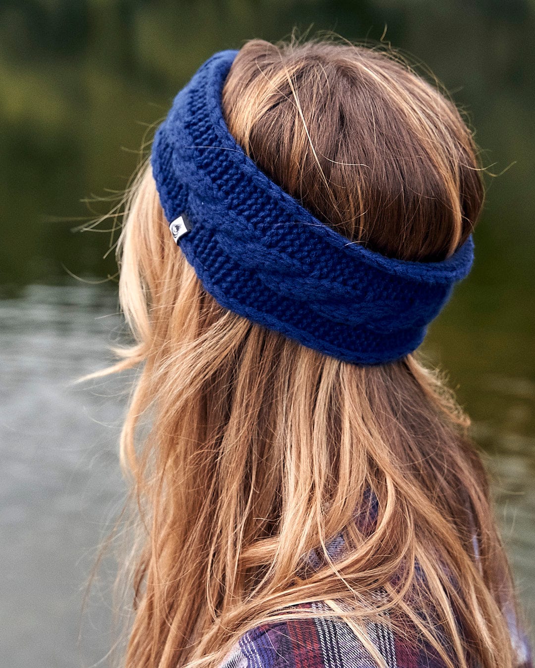 A woman wearing a Saltrock Cable - Knitted Headband - Blue by a lake, showcasing both fashion and warmth.