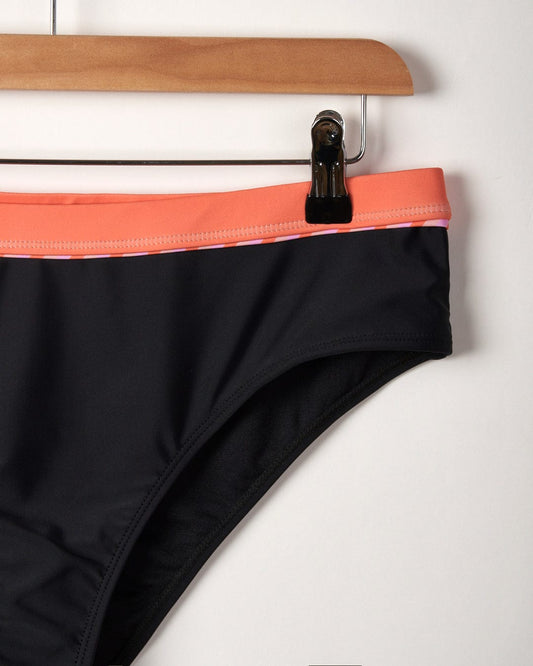 Close-up of Caeley - Recycled Womens Retro Bikini Bottoms in black made from polyester elastane with a peach-colored waistband hanging on a wooden hanger against a white background by Saltrock.