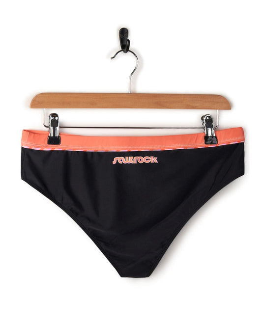 Caeley - Recycled Womens Retro Bikini Bottoms in Black from Saltrock with a coral waistband, crafted from polyester elastane and hanging on a wooden hanger against a white background.