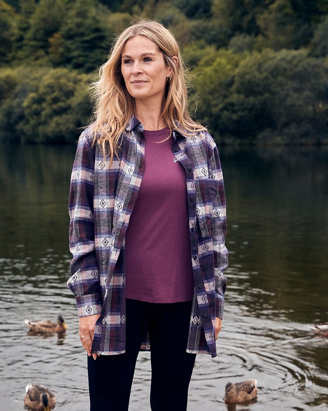 A woman wearing a Saltrock Camille - Womens Jaquard Check Boyfriend Shirt in Purple stands by a lake with ducks in the background.
