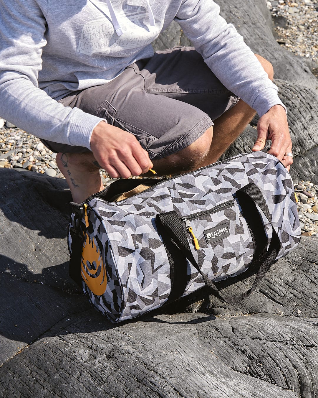 A man sitting on rocks with a Saltrock Camo Balboa - Hold-All Bag - Dark Grey.