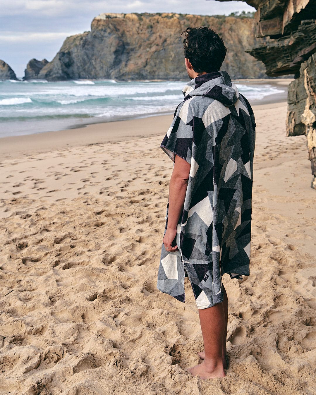 A man standing on a beach with a Saltrock Geo Camo - Changing Towel - Grey.