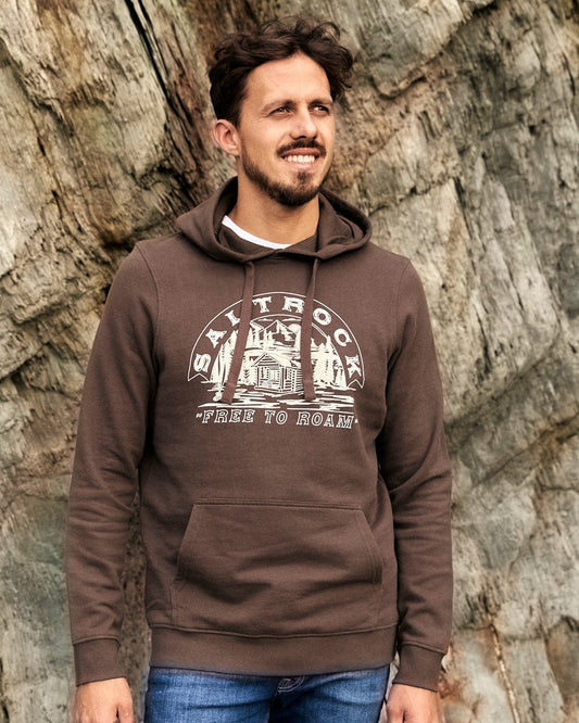 A man wearing a Camping Cabin - Mens Pop Hoodie in Brown by Saltrock, with a drawstring hood, standing in front of a rock.
