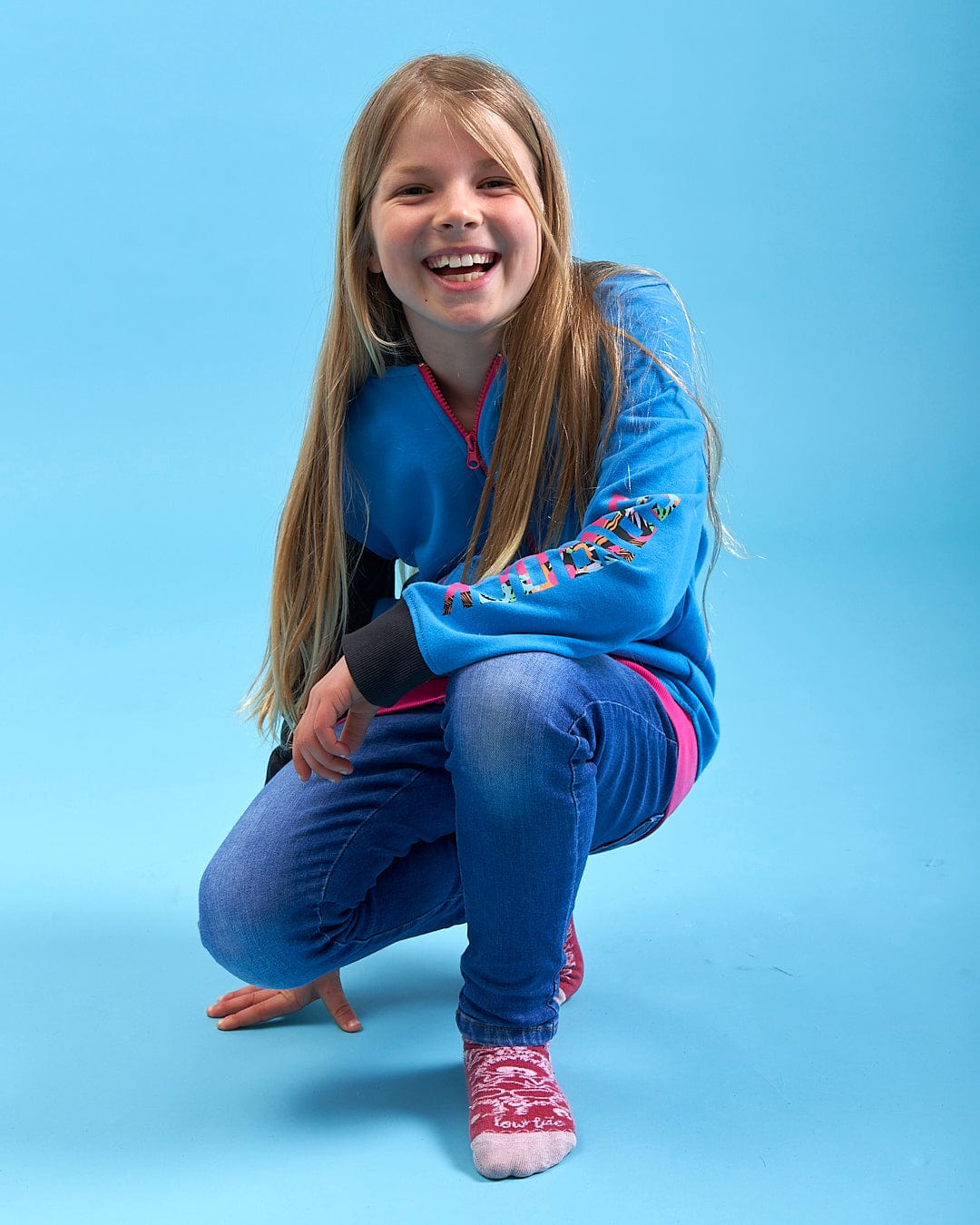 A girl wearing a Cara - Girls Zip Hoodie - Blue from Saltrock, squatting on a blue background.