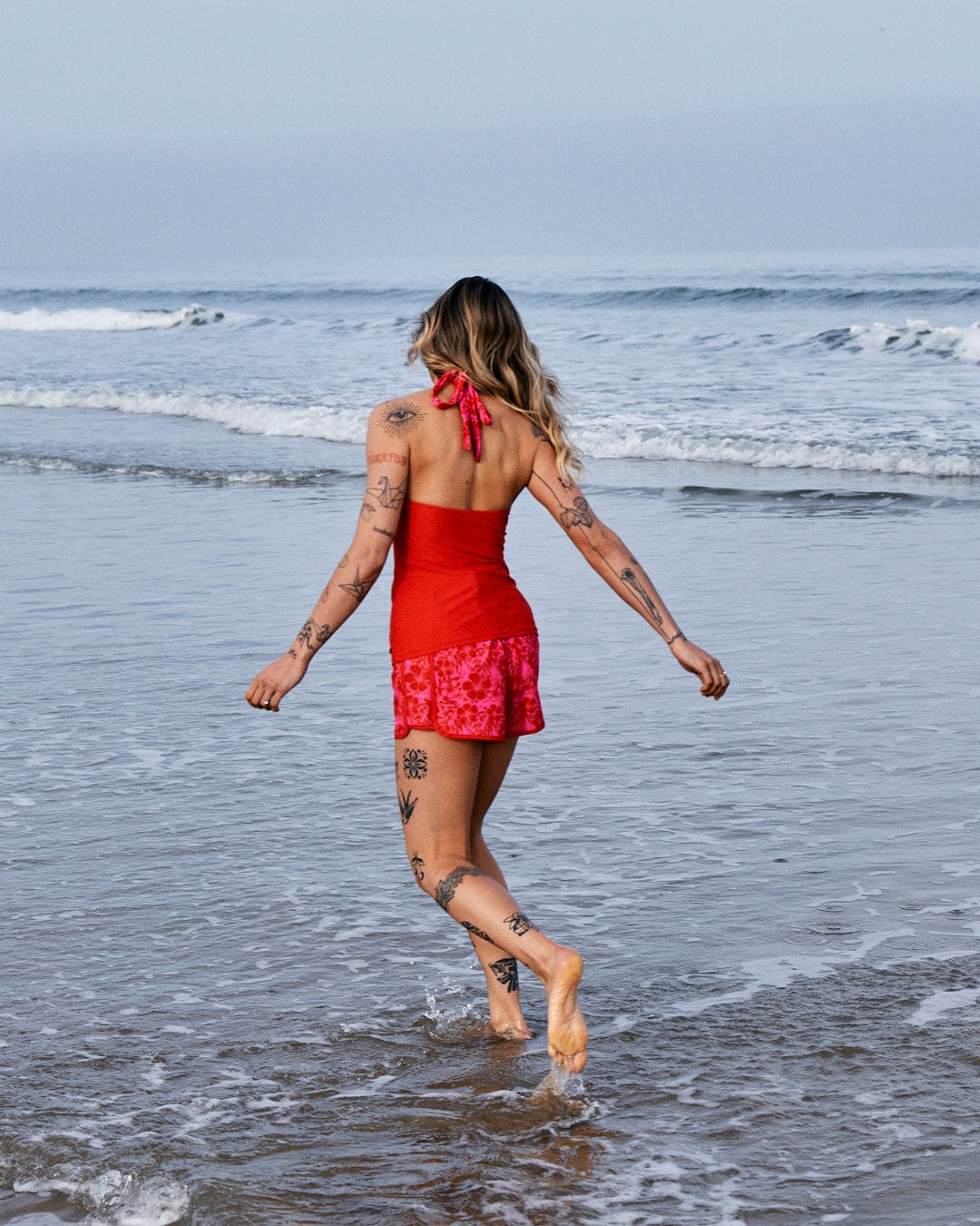 Woman with tattoos walking into the ocean waves, wearing a red dress made of recycled fibers and a Carly Hibiscus - Recycled Womens Halter Neck Bikini Top - Pink scarf in her hair by Saltrock.