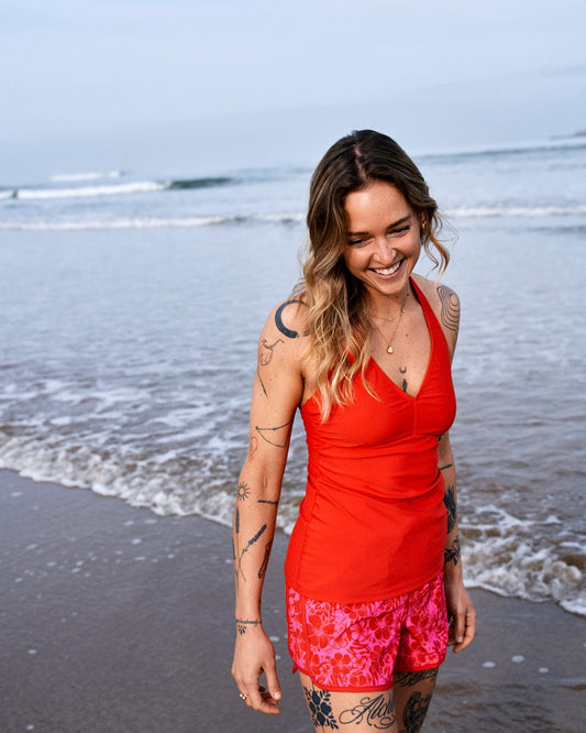 A smiling woman with tattoos walks along a beach, wearing a Saltrock Carly Hibiscus - Recycled Womens Halter Neck Bikini Top in Pink and patterned shorts, with waves in the background.