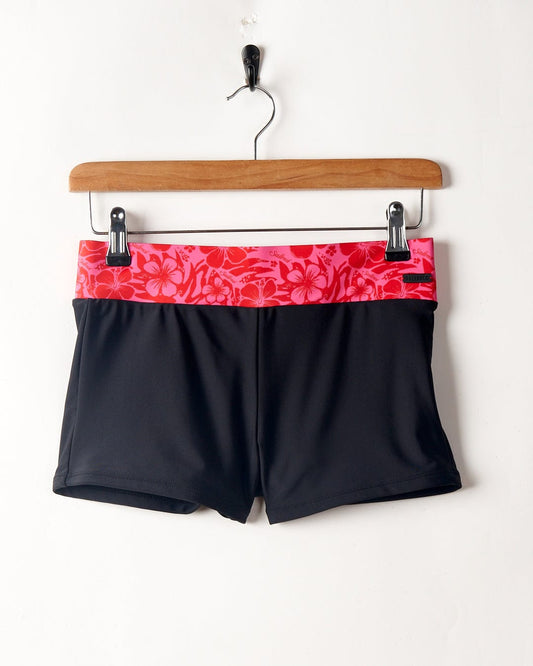 A pair of black Cassie Hibiscus - Recycled Bikini Bottoms with a red waistband hanging on a wooden hanger against a white wall. Brand: Saltrock.