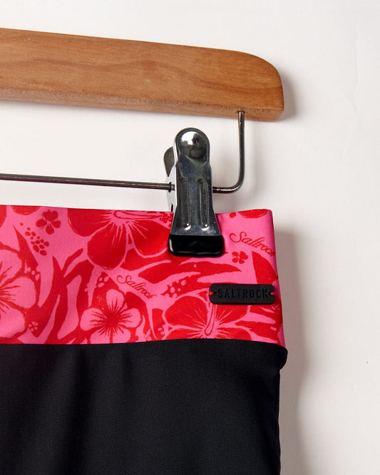 Close-up of a Cassie Hibiscus - Recycled Bikini Bottoms - Black/Pink saltrock skirt with a black hem, held by a wooden hanger with a metal clip.