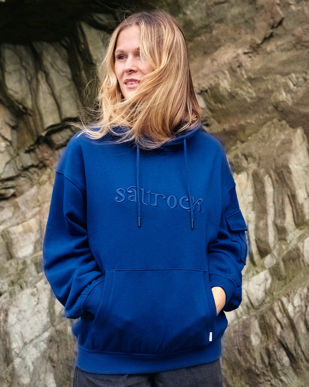 A woman wearing a blue Celeste - Womens Pop Hoodie - Blue with embroidered Saltrock branding, in front of rocks.