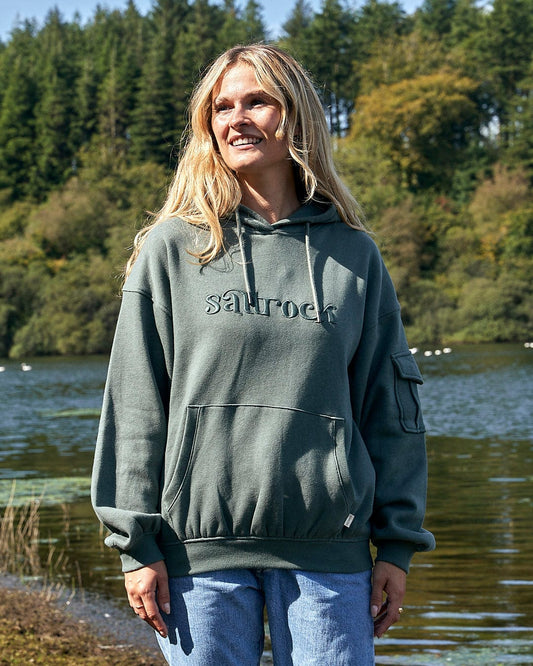 A woman wearing a Saltrock Celeste - Womens Pop Hoodie - Dark Green with drawstring hood stands next to a lake.