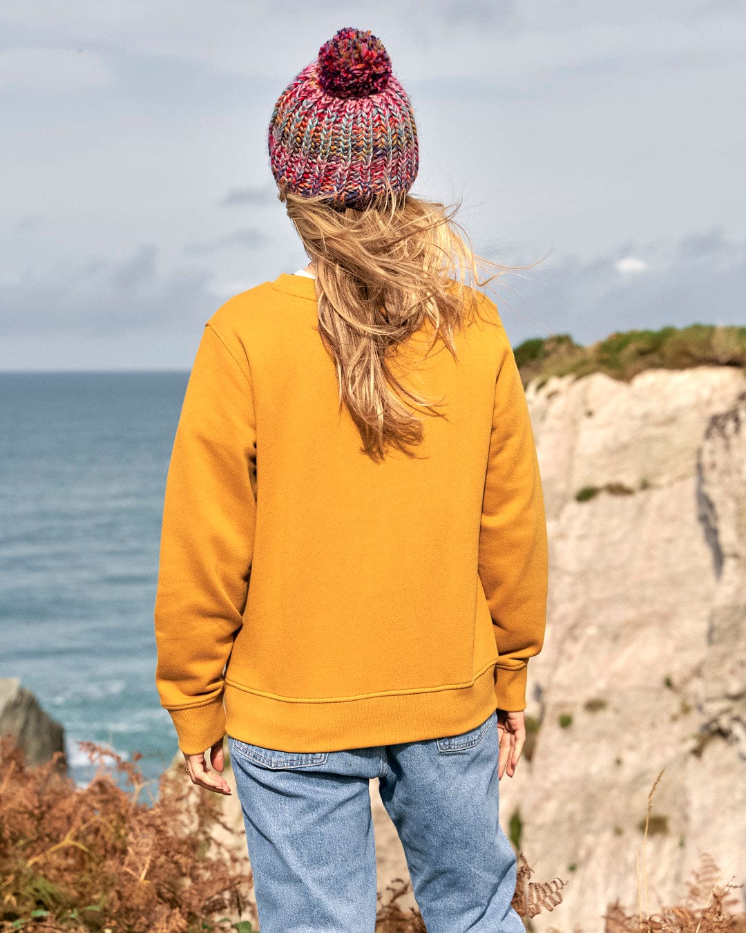 A woman wearing a yellow Saltrock sweatshirt, adorned with Saltrock branding, standing on a cliff overlooking the ocean.