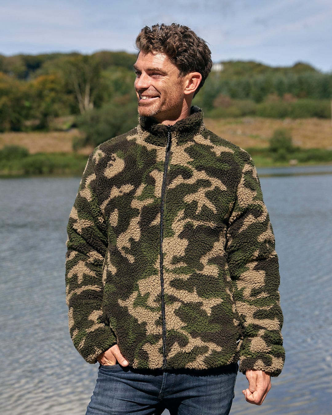 A man wearing a Saltrock Cirrus - Quilted Reversible Jacket - Dark Green standing by a lake.