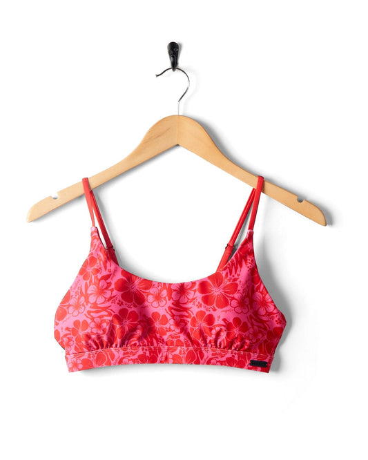 A Cleo Hibiscus - Recycled Bikini Top - Pink by Saltrock, with adjustable thin straps, hung on a wooden hanger against a white background.
