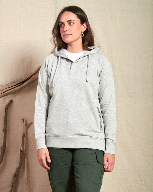 A woman wearing the Saltrock Cliffden Women's Pop Hoodie in grey and green cargo pants stands indoors against a beige fabric backdrop with wooden branches.