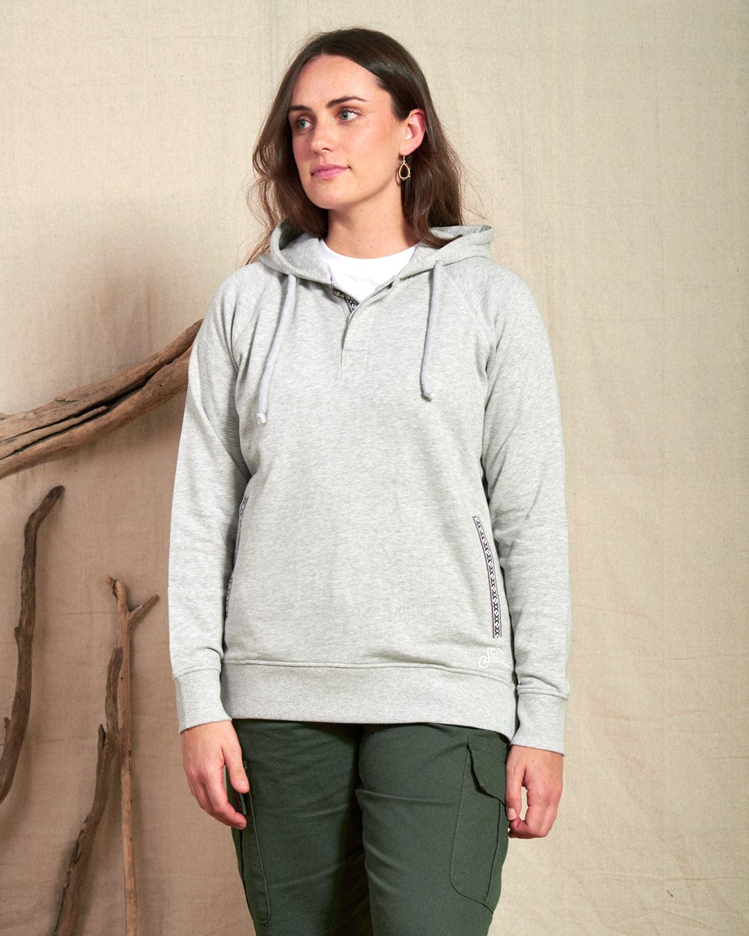 A woman wearing the Saltrock Cliffden Women's Pop Hoodie in grey and green cargo pants stands indoors against a beige fabric backdrop with wooden branches.