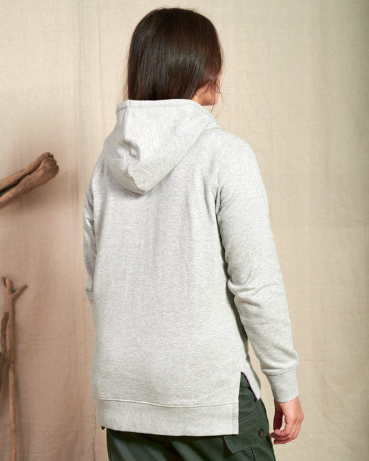 Person facing away from the camera, wearing a light gray Saltrock Cliffden Womens Pop Hoodie and dark green pants, standing indoors against a beige background.