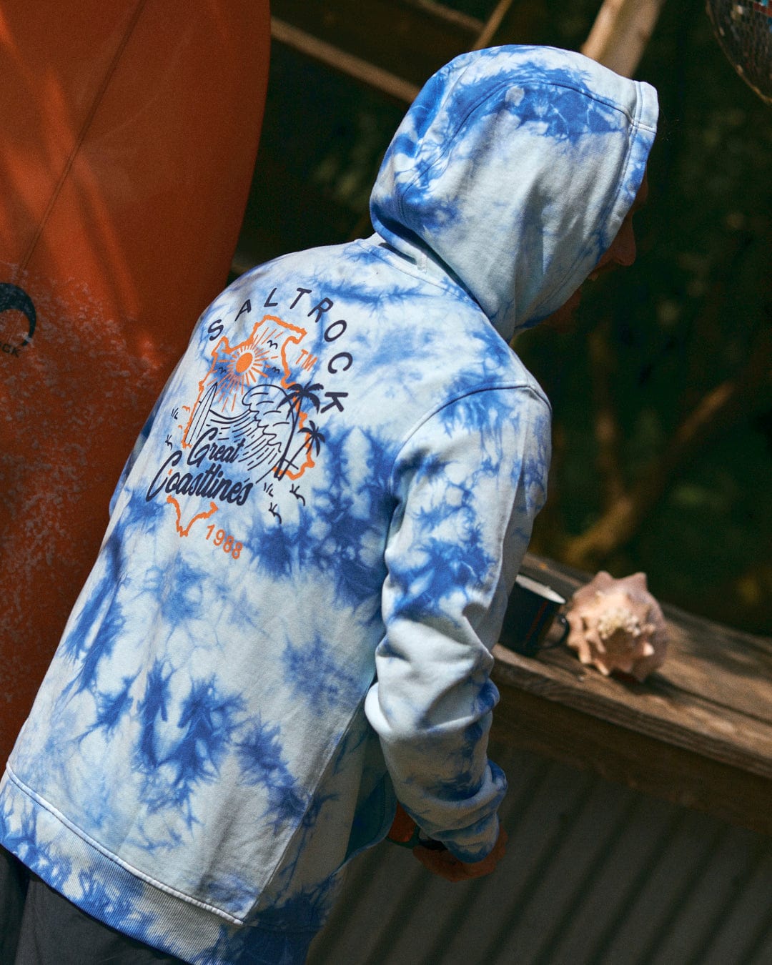 A person is standing outdoors, wearing the Saltrock Coastline Men's Tie Dye Pop Hoodie in blue. The hoodie features a stylish tie-dye design and the word "Togo" on the back, and is made from 100% cotton.