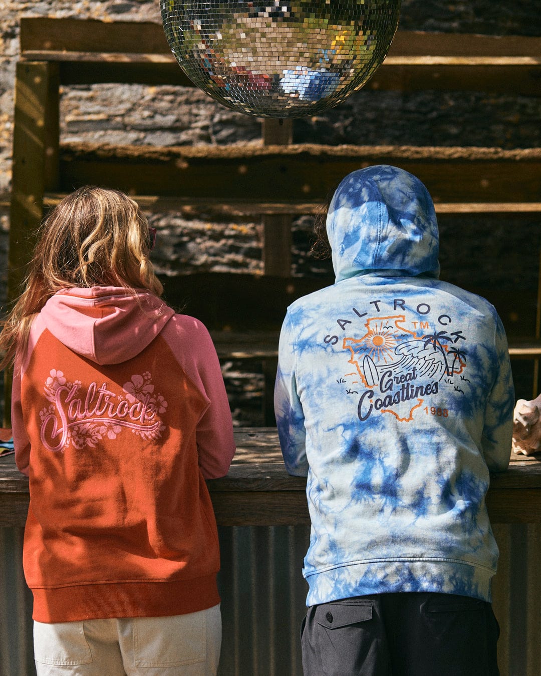 Two individuals wearing Saltrock hoodies, one in orange and the other in the Saltrock Coastline - Mens Tie Dye Pop Hoodie - Blue made of 100% cotton, stand with their backs to the camera under a reflective disco ball.