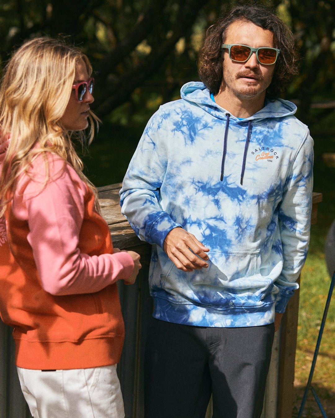 Two people wearing sunglasses and hoodies stand outdoors near a wooden structure; the person on the left is wearing a pink hoodie, while the person on the right sports the Saltrock Coastline Men's Tie Dye Pop Hoodie in Blue. Both hoodies are made from 100% cotton.