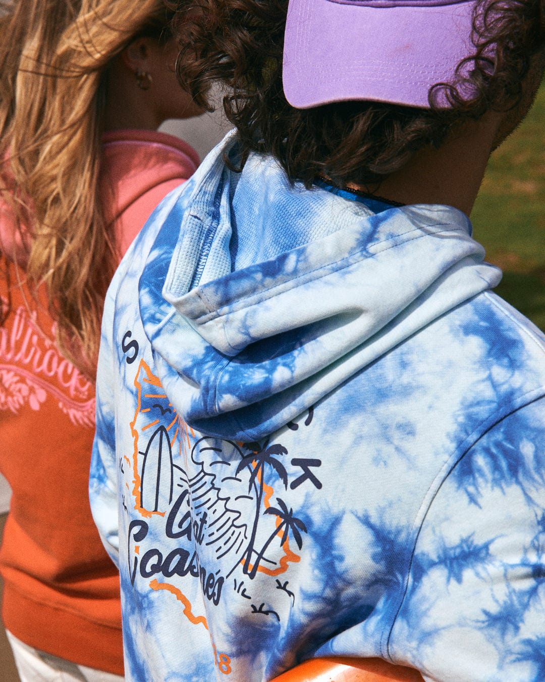 Two people are shown from behind, wearing casual clothing. One person sports a purple hat and the Saltrock Coastline - Mens Tie Dye Pop Hoodie in blue with a California-themed graphic. The other wears a red-orange sweatshirt.