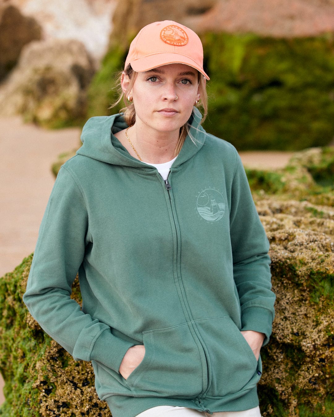 A young woman wearing a green hoodie and a Saltrock Cold Water Club cap in orange with a curved visor stands outdoors with rocky formations in the background.