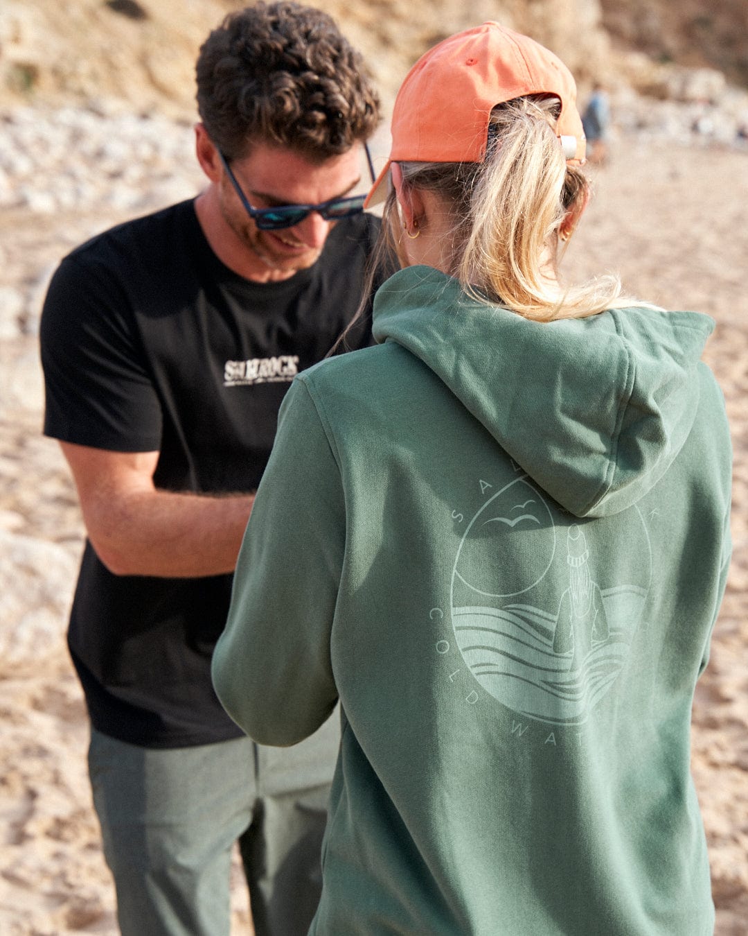 Two people engaged in a conversation on a sandy beach, wearing Coldwater Club - Ladies Zip Hoodie - Green Saltrock shirts.