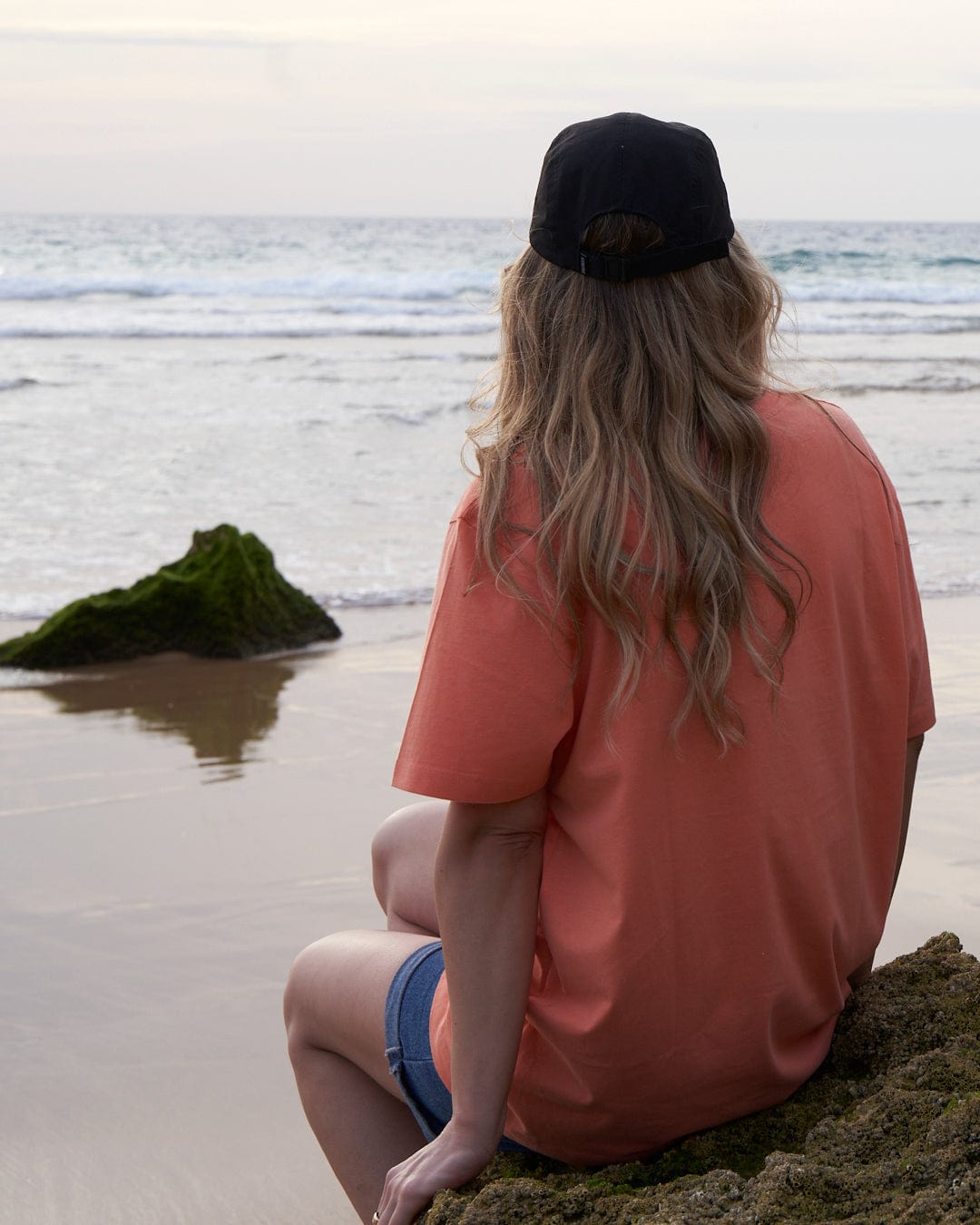 A person with long hair, wearing a Saltrock Gaitor 5 Panel UPF Cap - Black with an adjustable strap and an orange shirt, sits on a rock and gazes out at the ocean waves.
