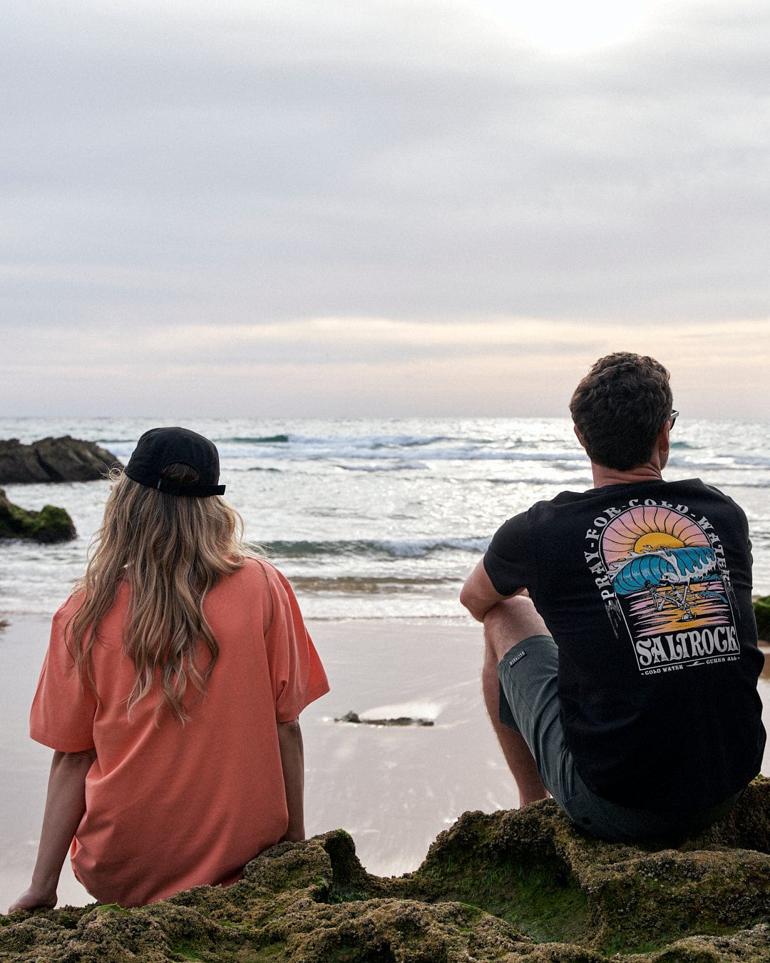 Two people sitting on rocks at the beach wearing Saltrock Coldwater Club Womens Short Sleeve T-Shirts in Peach, looking at the ocean.