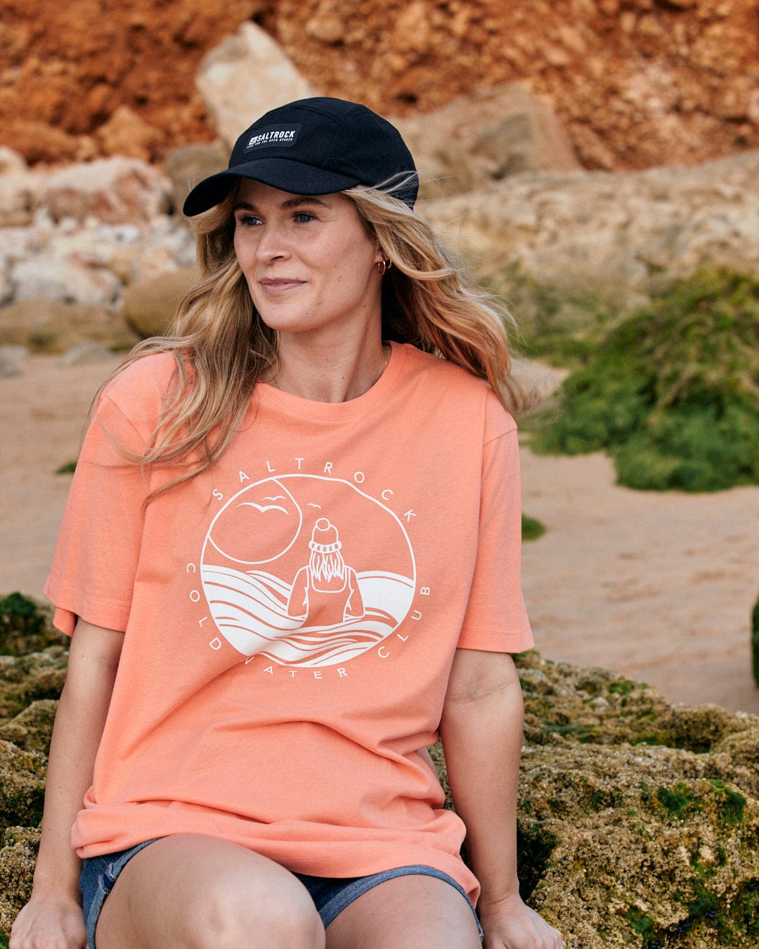 A woman wearing a Saltrock Coldwater Club - Womens Short Sleeve T-Shirt in Peach sitting on rocks.