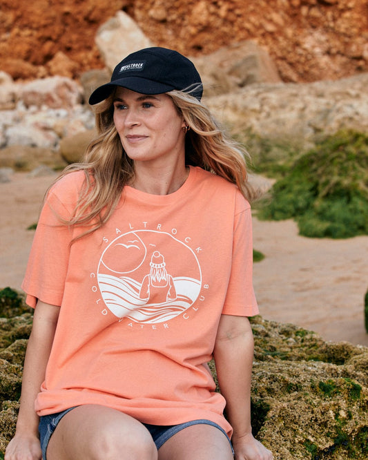 A woman in a water-resistant coral t-shirt and Saltrock Gaitor 5 Panel UPF Cap in black with an adjustable strap sits on a rocky beach, looking thoughtfully to the side.