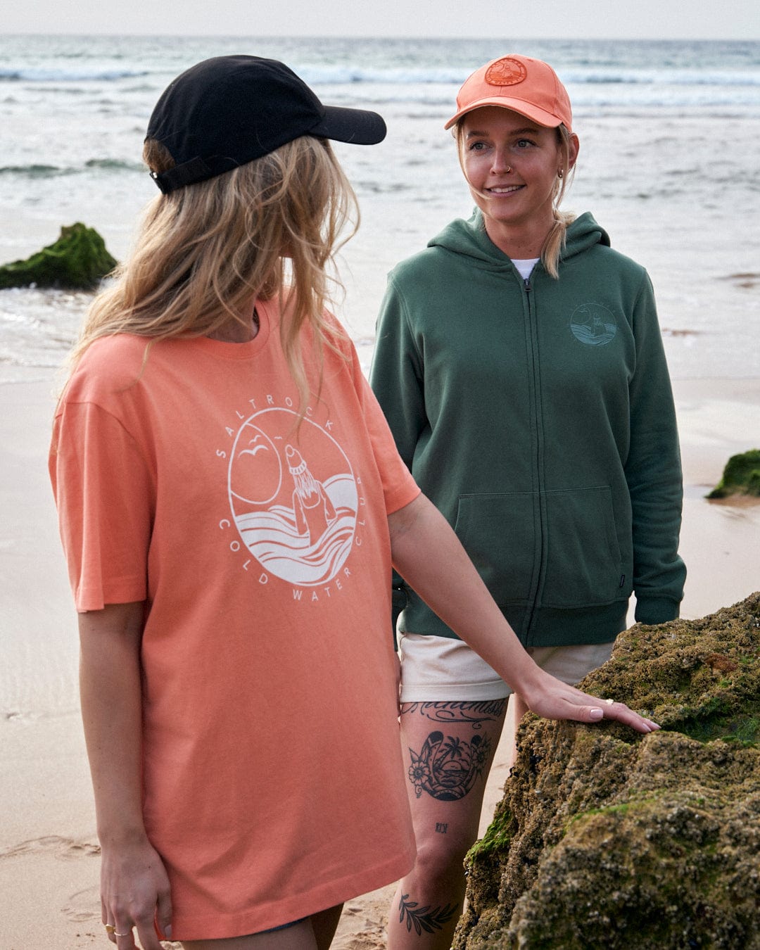 Two women wearing casual clothing with UPF protection and hats stand near a rocky beach. The woman on the left is facing away from the camera, revealing a black Gaitor 5 Panel UPF Cap with a graphic design on the back by Saltrock.