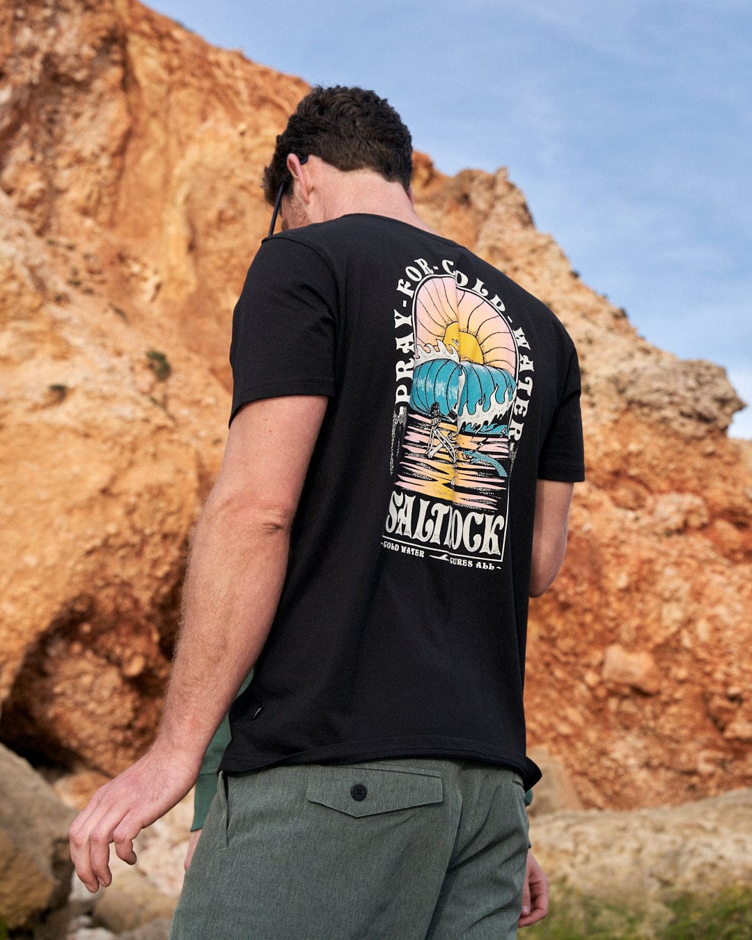 A man wearing a black t-shirt with Saltrock branding is standing on a rock.