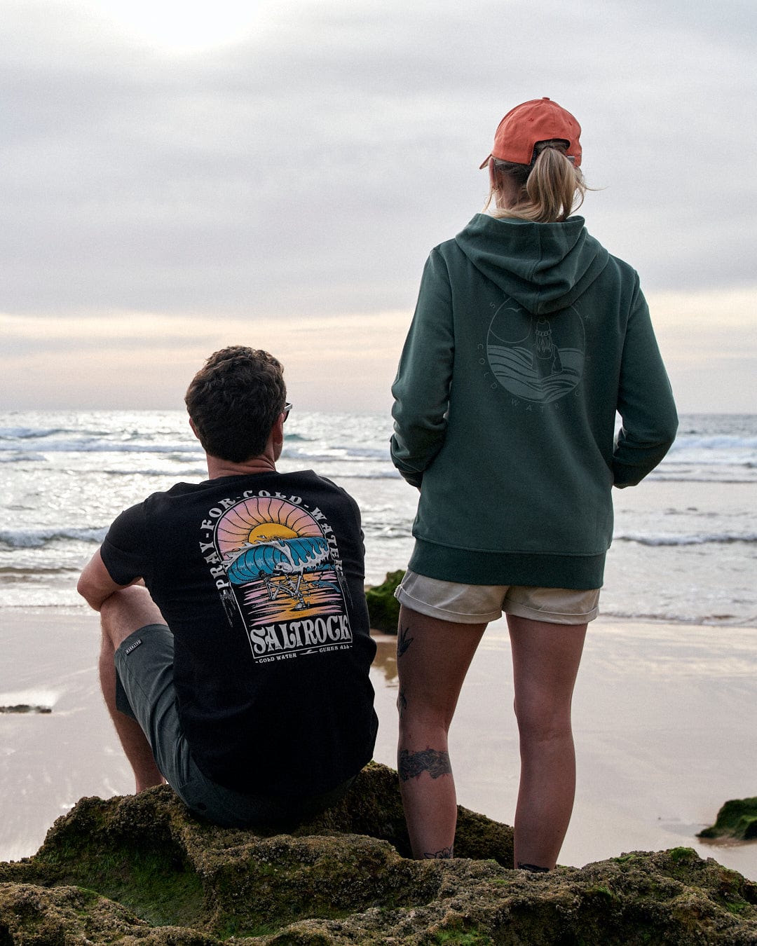 A man and woman sitting on Saltrock-branded Cold Water - Mens Short Sleeve T-Shirt - Black at the beach.