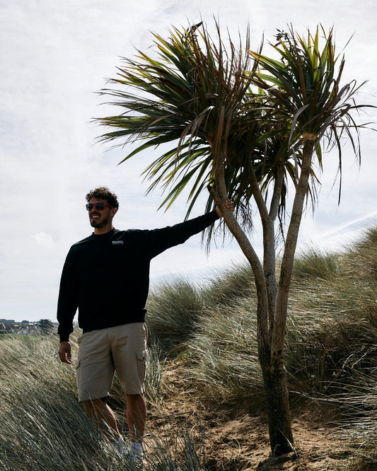 Man in sunglasses and casual Saltrock Cold Water - Mens Sweat - Black attire standing next to a palm tree on a sandy area with dunes in the background.