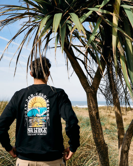 Person wearing a Saltrock Cold Water - Mens Sweat - Black sweatshirt made of 100% Cotton standing by a palm tree, facing a beach in the background.