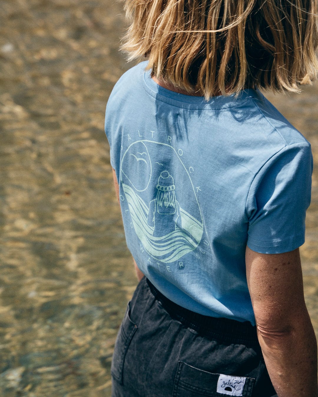 A person with shoulder-length blonde hair wears the Saltrock "Cold Water Club" Women's T-Shirt in blue, featuring a graphic on the back, while standing next to clear water.