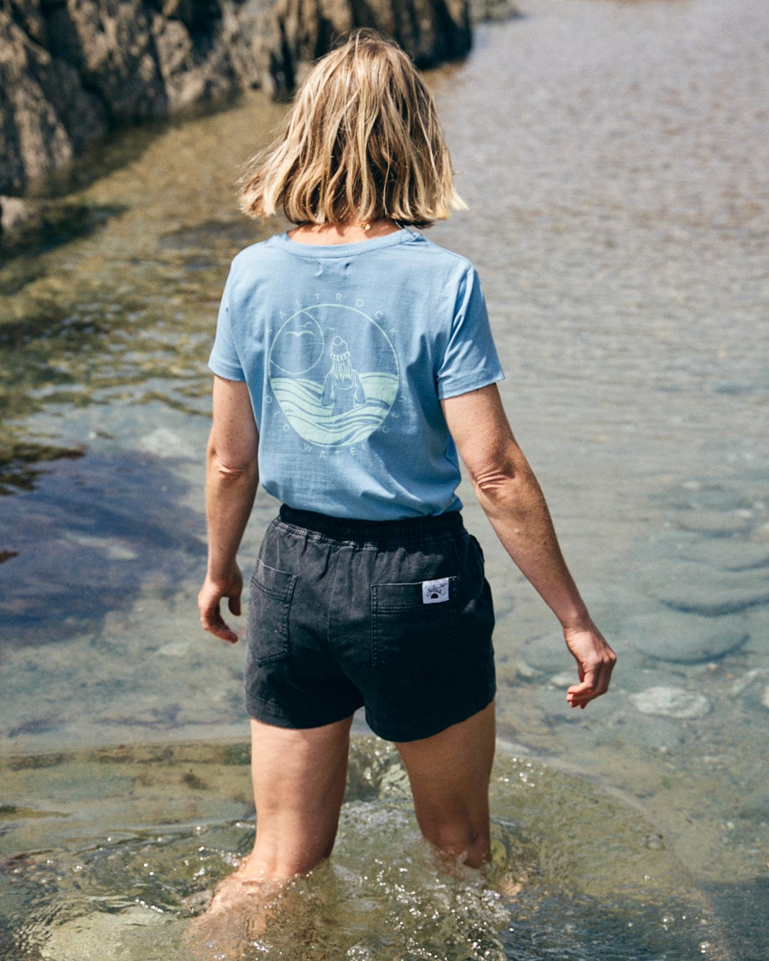 A person with short blond hair stands in shallow water near rocks, dressed in a Saltrock Cold Water Club women's blue t-shirt and dark shorts.