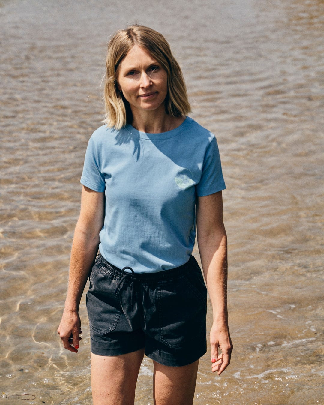 A woman with shoulder-length blonde hair, wearing a Saltrock Cold Water Club - Womens T-Shirt in blue and black shorts, stands in shallow water.