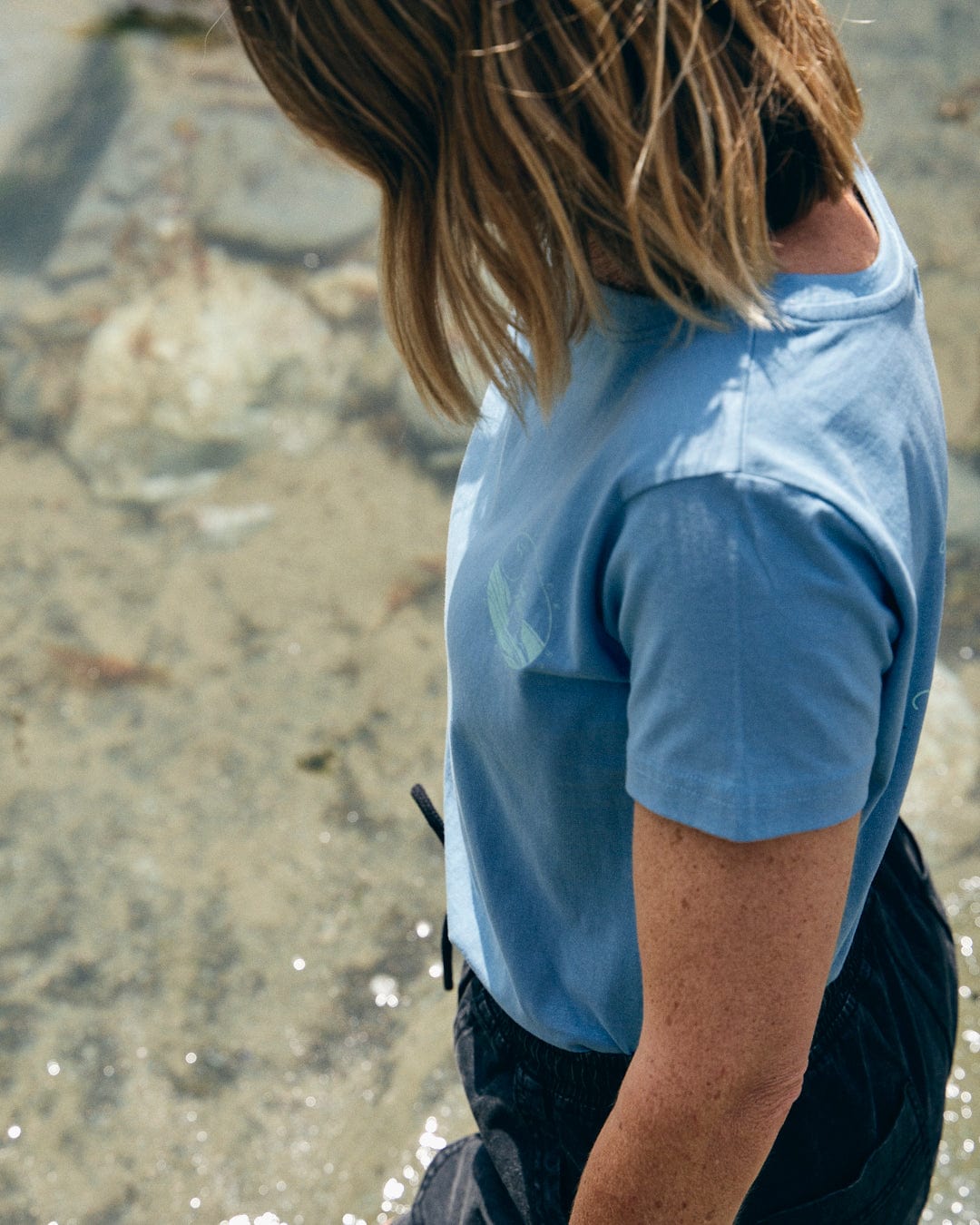 A person wearing the Saltrock "Cold Water Club" Women's T-Shirt in blue and dark pants stands on the rocky shore, looking down at the water.