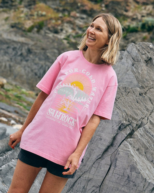 A person with shoulder-length hair is smiling while standing on rocky terrain, wearing a **Saltrock Cold Water women's short sleeve T-shirt in pink**, made of 100% cotton with a graphic design, paired with dark shorts.