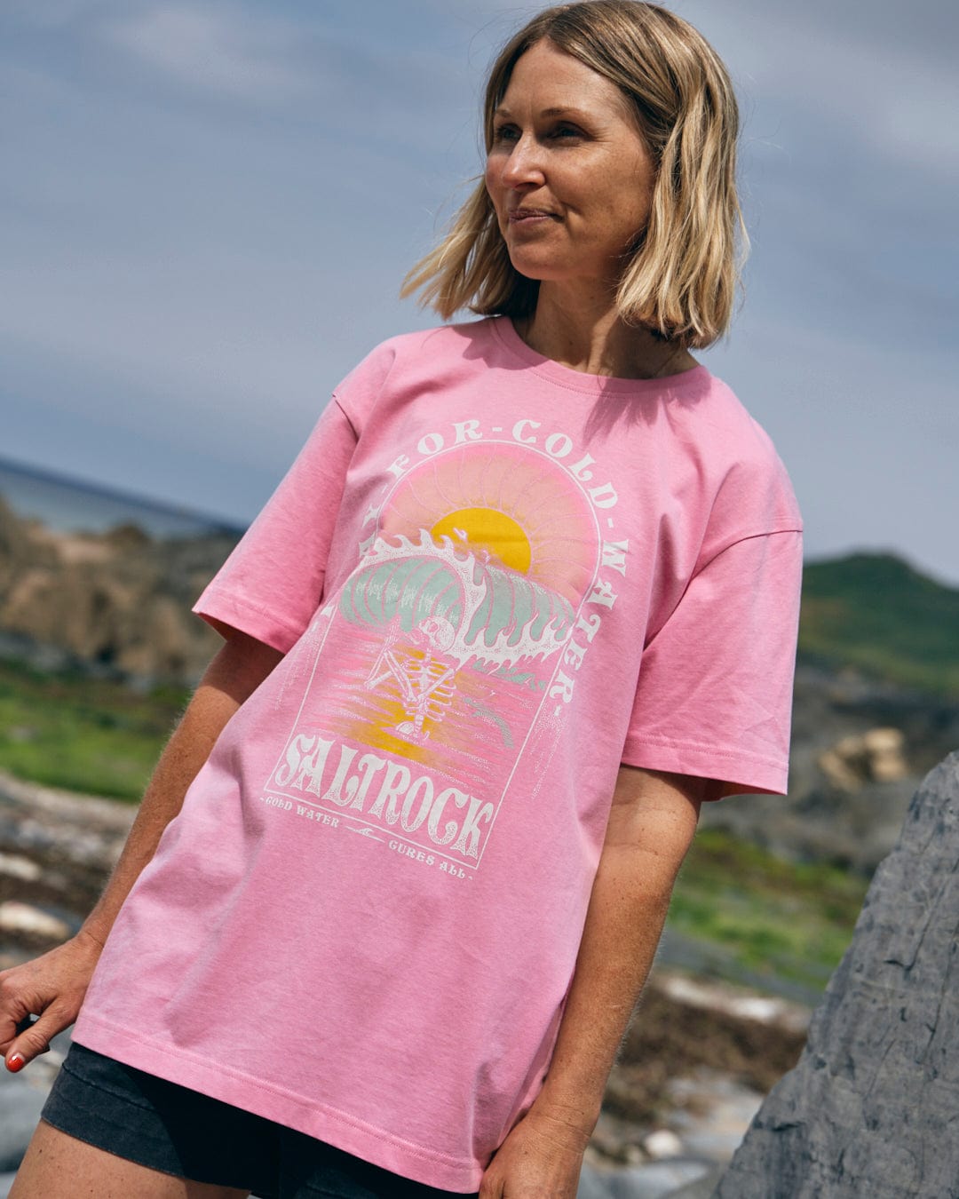 A woman wearing a pink "Cold Water - Women's Short Sleeve T-Shirt" from Saltrock stands outdoors near rocky terrain and greenery. She has blonde hair and is looking to the side.