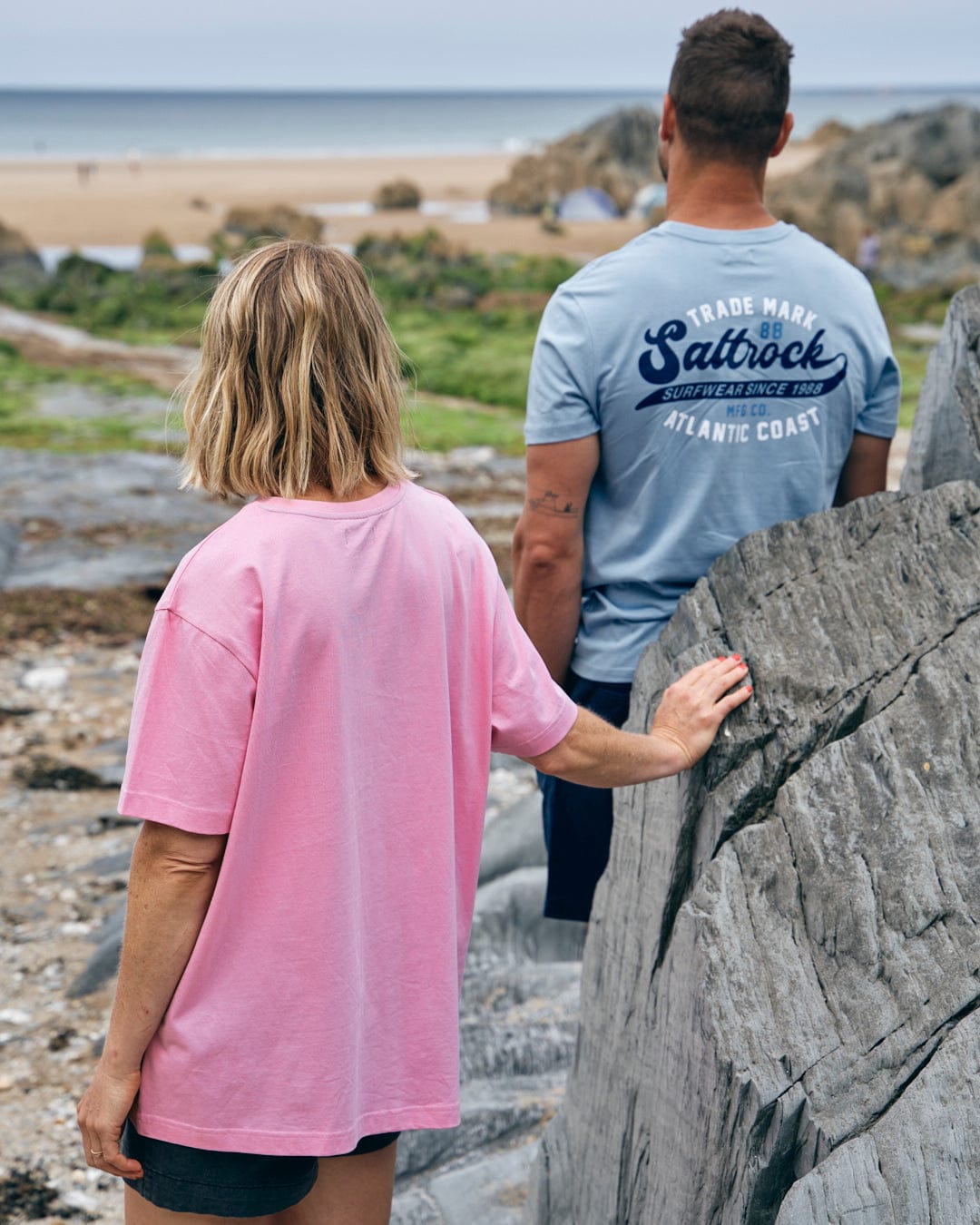 Two people stand on rocky terrain at the beach. The person in the foreground wears a "Cold Water - Womens Short Sleeve T-Shirt" in pink from Saltrock, while the person in the background sports a blue shirt with the "Saltrock" text. The ocean and sand are visible behind them.