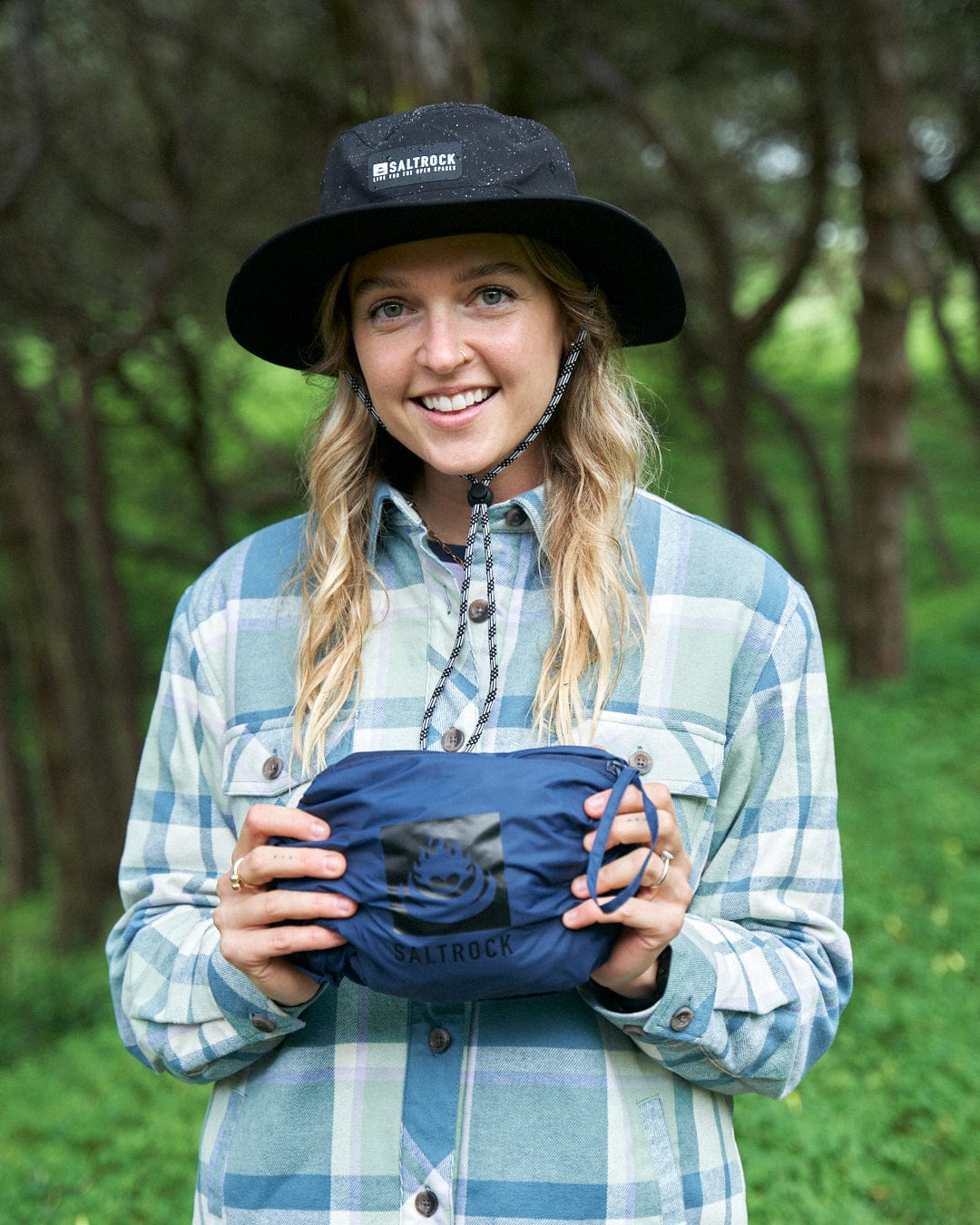 A smiling young woman in a plaid shirt and Saltrock waterproof jacket holds a Cooper - Womens Packable Waterproof Jacket - Blue bag in a lush green forest setting.