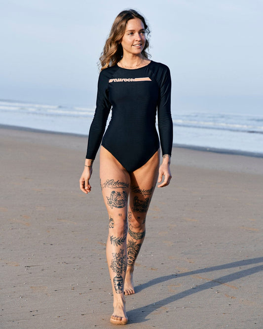 Woman with tattoos wearing a black Saltrock Cora Retro long-sleeve swimsuit made from recycled fibers, walking on a beach.