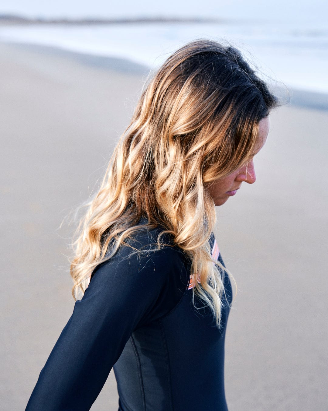 Profile view of a woman with blonde wavy hair, wearing a Saltrock Cora Retro - Recycled Womens Long Sleeve Swimsuit in Dark Grey, standing on a beach.