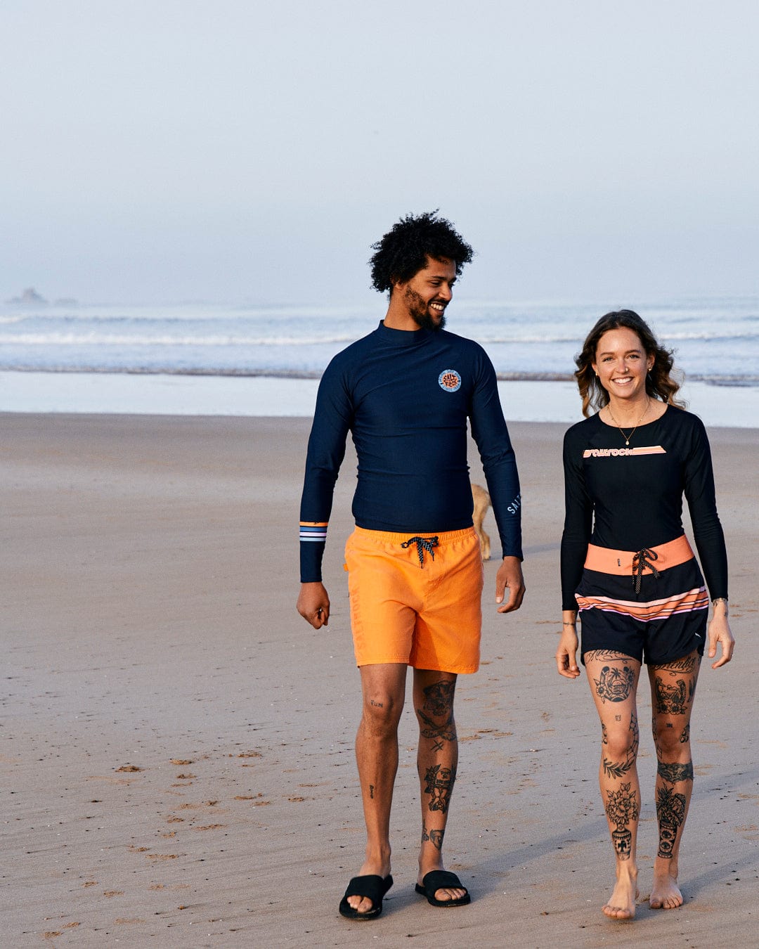 A man and a woman walking on a beach, both wearing Saltrock Cora Retro - Recycled long sleeve swimsuits in dark grey, smiling and looking at each other.