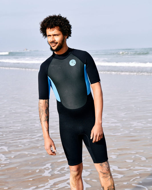 A man in a black and blue short-sleeve wetsuit stands on a beach with waves in the background. The Saltrock Core - Mens 3/2 Shortie Wetsuit - Black/Blue accentuates his curly hair and tattoos on his arms.