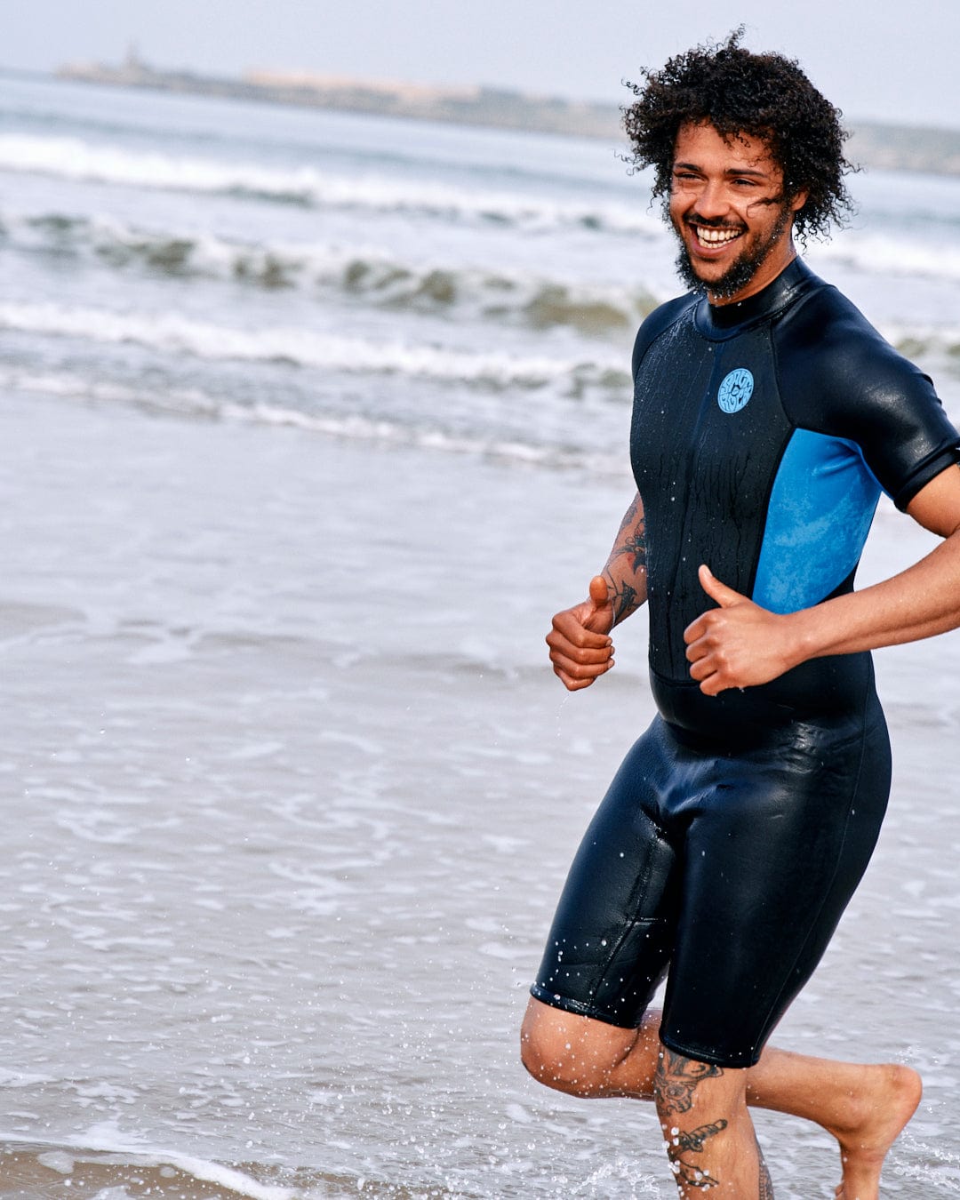 A joyful man in a Saltrock neoprene Core - Men's 3/2 Shortie Wetsuit - Black/blue running on the beach, with waves in the background. He has curly hair and tattoos on his arm.