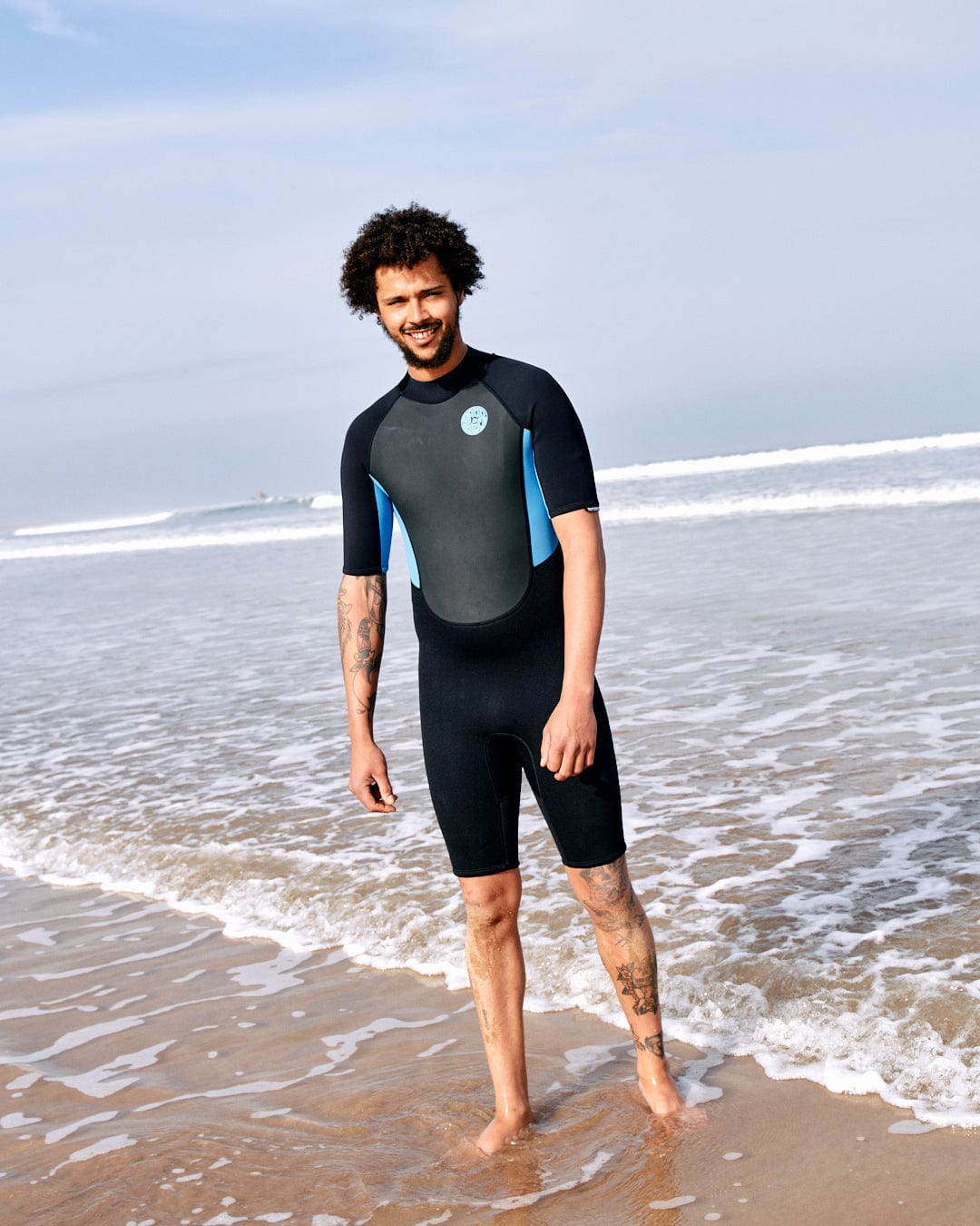 A man in a Core - Men's 3/2 Shortie Wetsuit in Black/blue by Saltrock standing on a sandy beach with waves in the background, smiling at the camera.